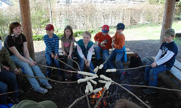 Eine Liz-Mitarbeiterin sitzt mit einer Gruppe Kinder am Lagerfeuer und grillt Stockbrot.