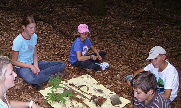 Eine Liz Mitarbeiterin sitzt mit 4 Kindern im Kreis und betrachten Natur-Fundstücke aus dem Wald.