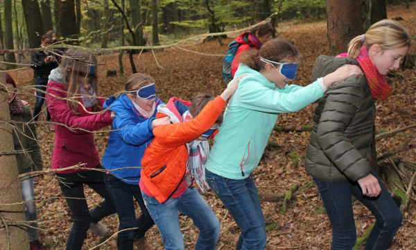 Eine Gruppe Kinder mit verbundenen Augen fassen sich von hinten an die Schulter und werden durch den Wald geführt.